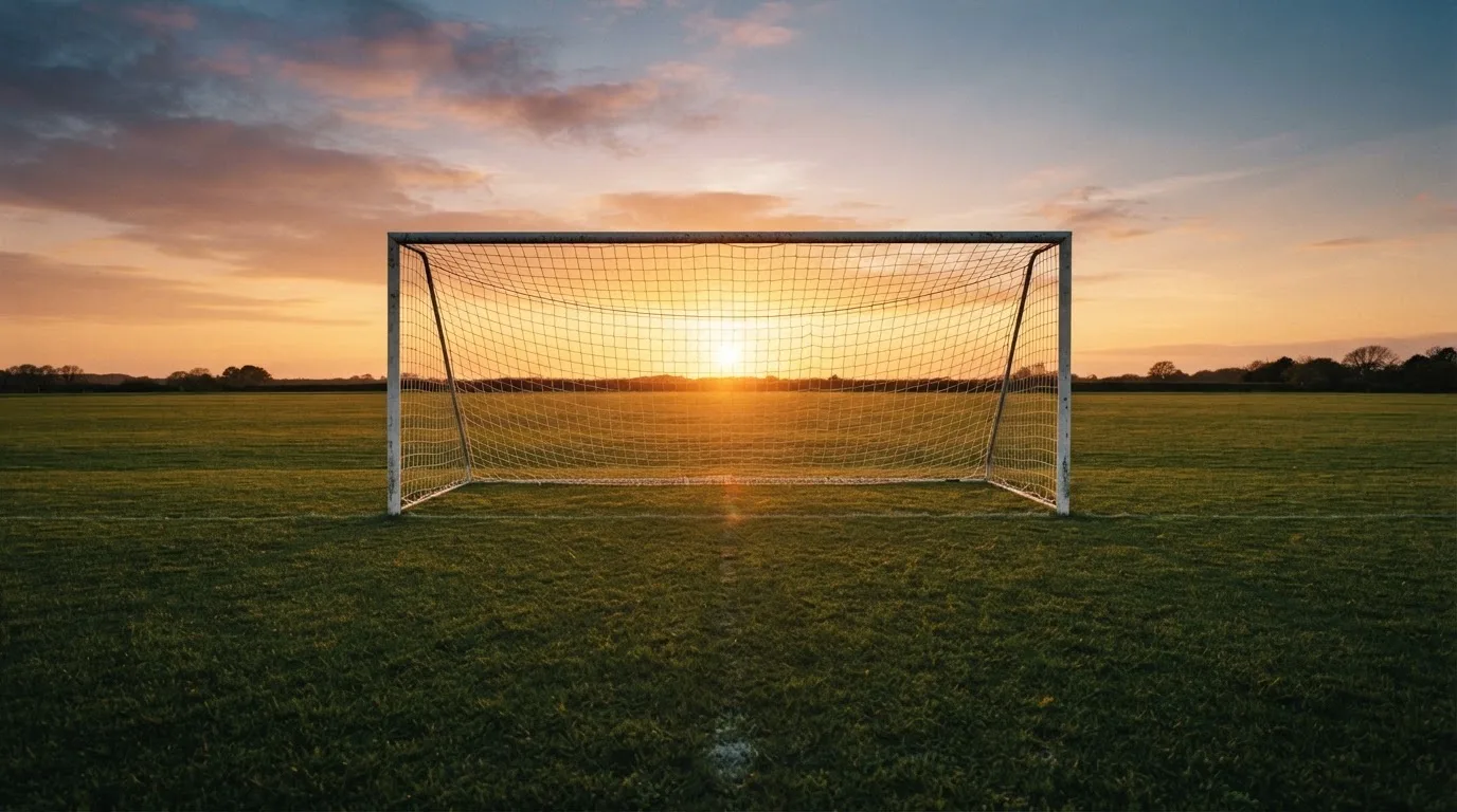 Ein Fußballtor mit Netz bei Sonnenuntergang auf einem leeren Platz, die Schatten der Torpfosten liegen lang auf dem Gras.