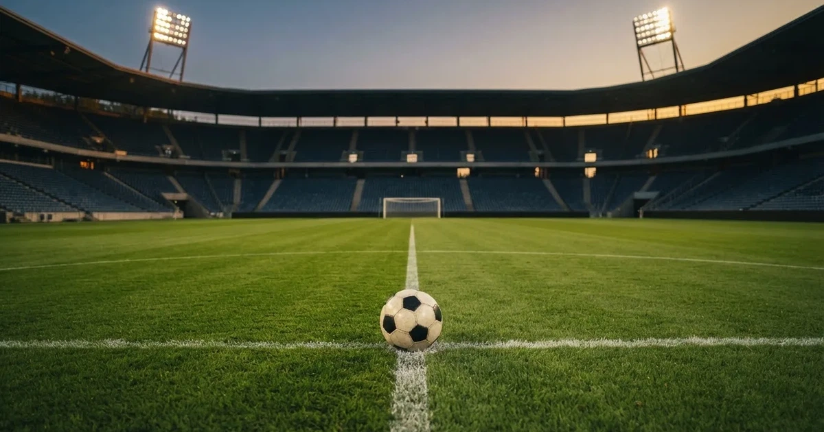 Ein Fußball liegt auf dem Anstoßpunkt eines leeren Rasenplatzes, im Hintergrund eine große Stadiontribüne im Abendlicht.