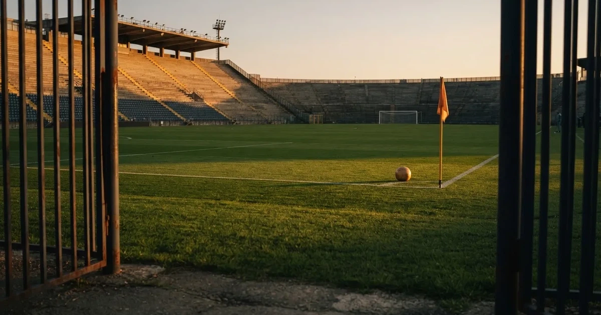 Blick durch ein Stadiontor auf einen grünen Rasen im Abendlicht, ein Ball ruht an der Eckfahne, die Tribünen sind leer.