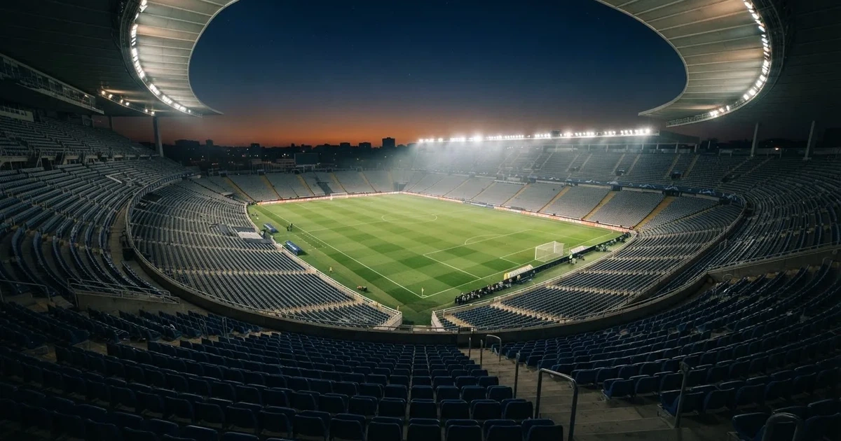 Leere Sitzreihen eines großen Stadions unter Flutlicht, dahinter die ferne Sicht auf den hell beleuchteten Rasen nach Sonnenuntergang.