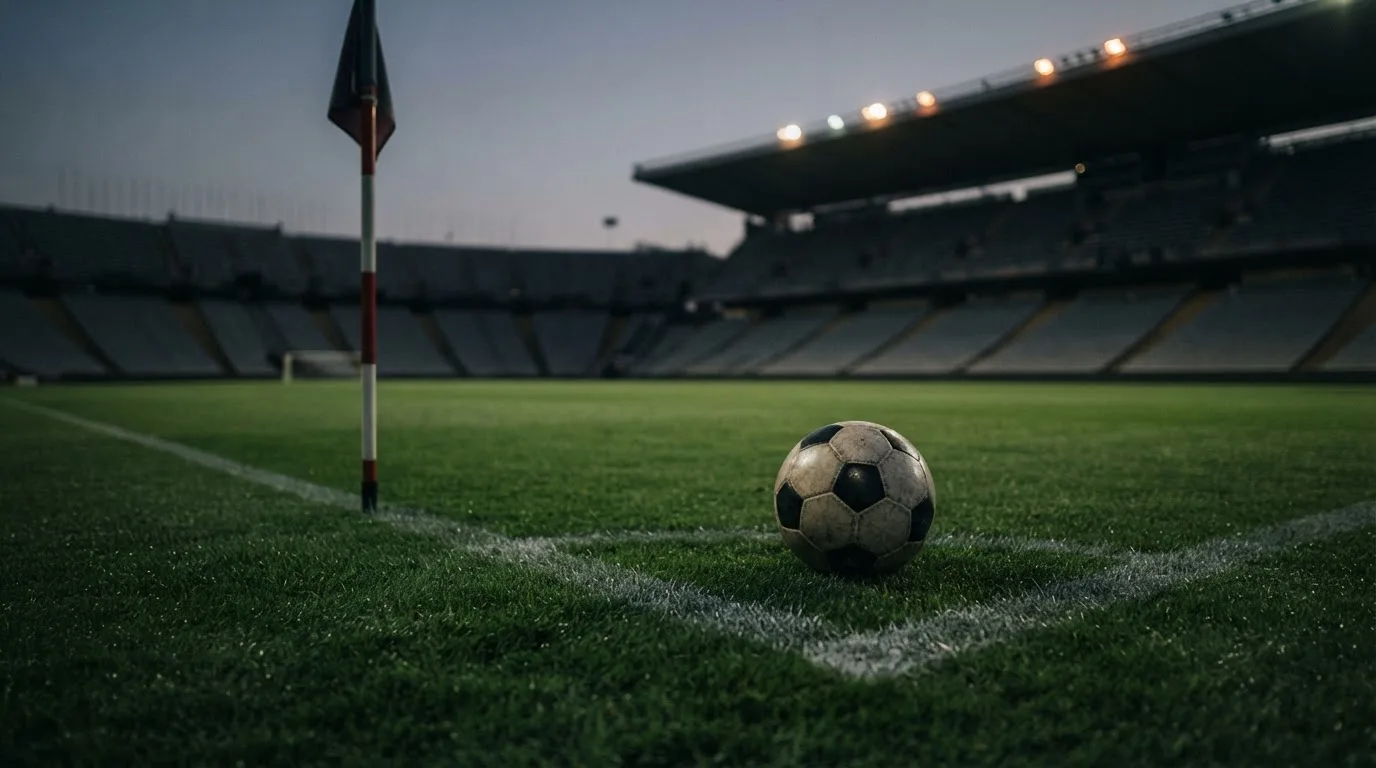 Ein Fußball liegt neben einem Eckfahnen-Pfosten auf einem Rasenplatz, im Hintergrund ein leeres Stadion in der Dämmerung.