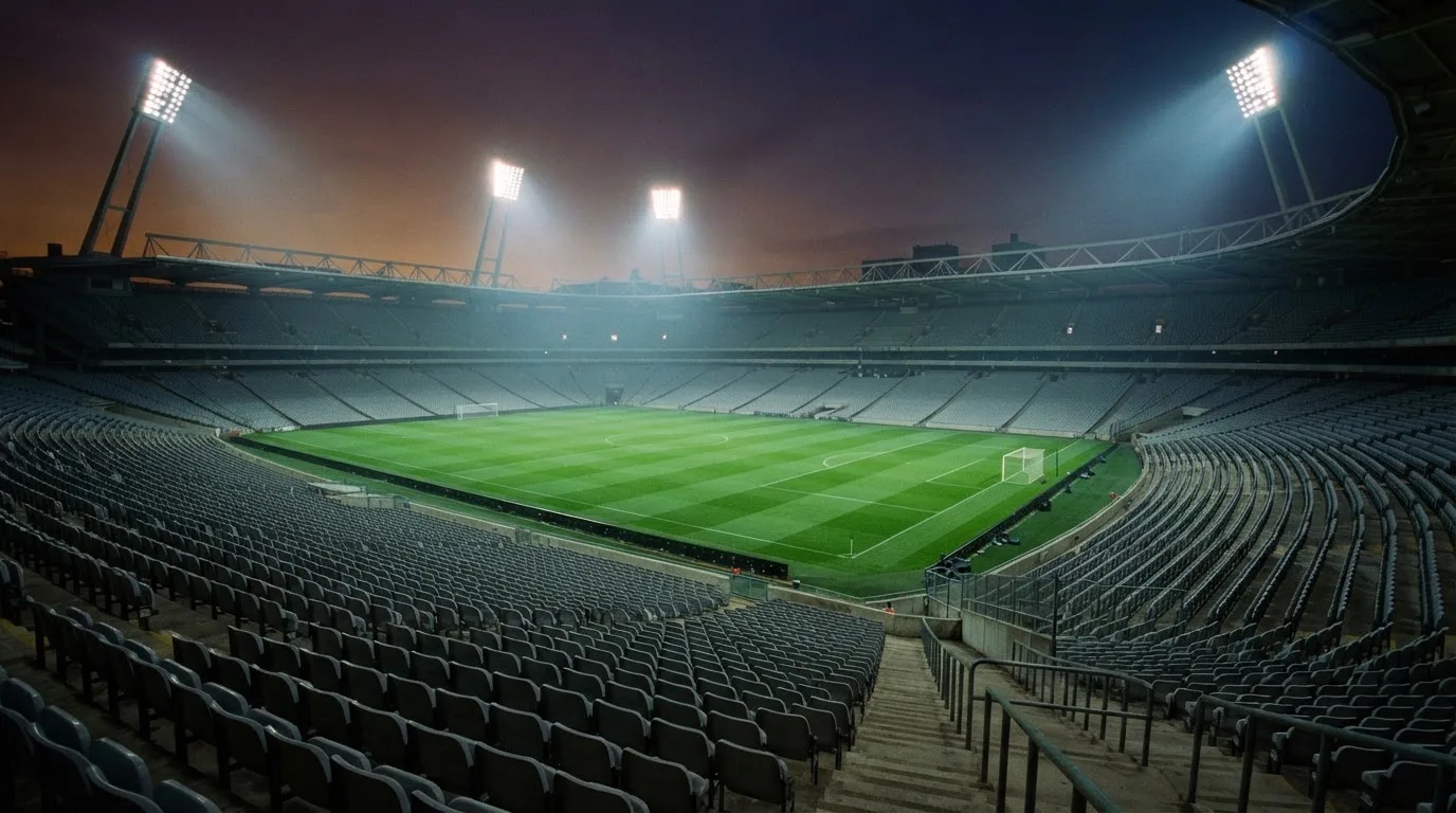 Eine leere Stadiontribüne unter Flutlicht aus der Ferne betrachtet, die Rasenfläche leuchtet grün in der Dämmerung.