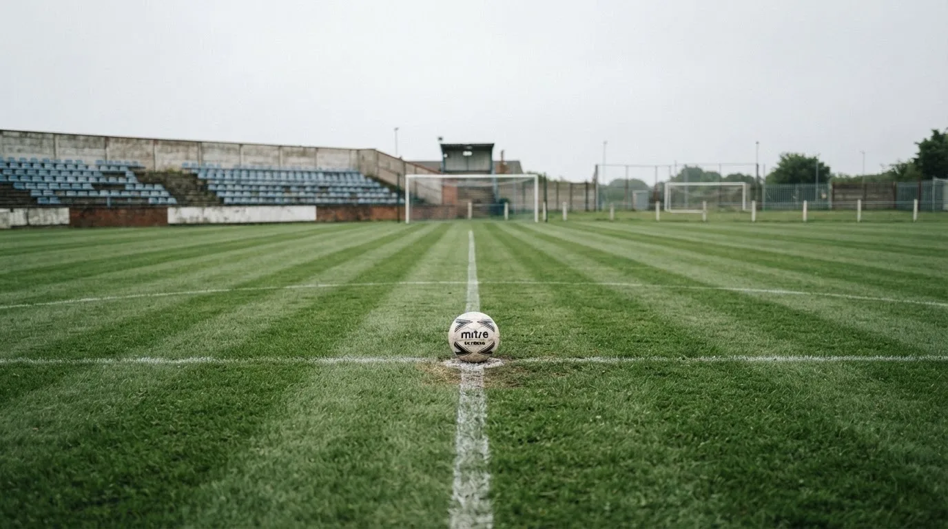 Ein Fußball liegt auf dem Anstoßpunkt eines leeren Rasenplatzes, im Hintergrund eine ruhige Tribüne unter bedecktem Himmel.