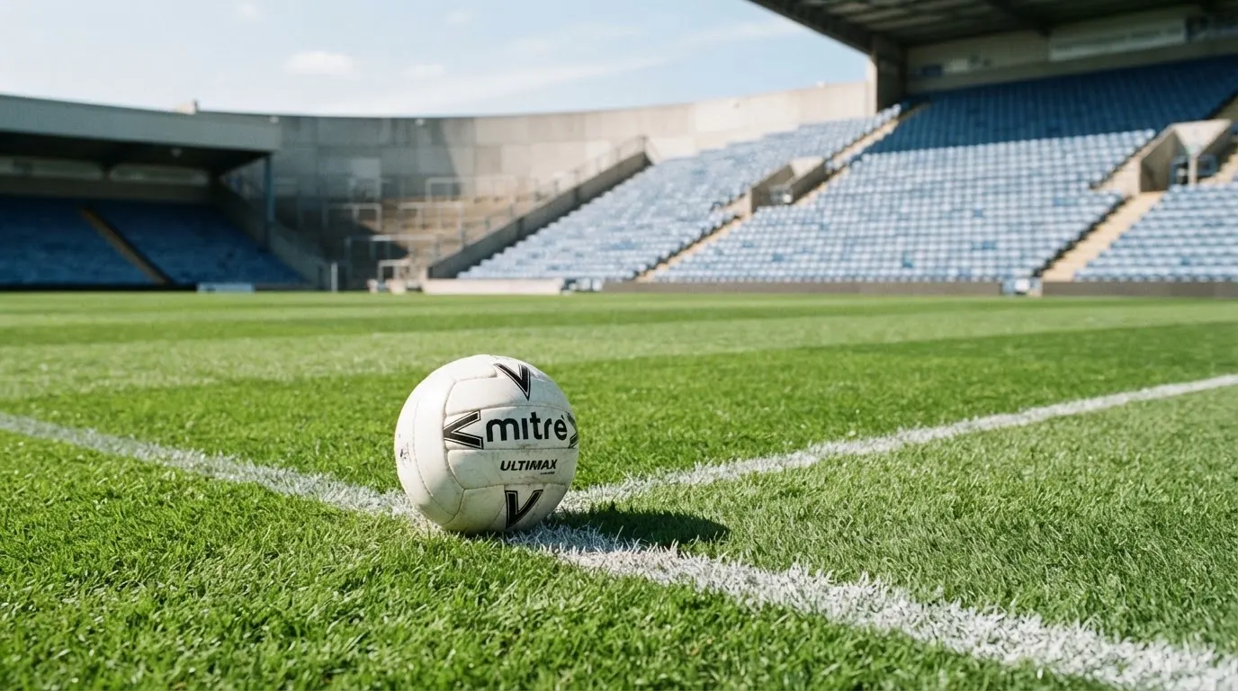 Ein Fußball liegt am Spielfeldrand eines Rasenplatzes, im Hintergrund eine leere Tribüne unter hellem Himmel.
