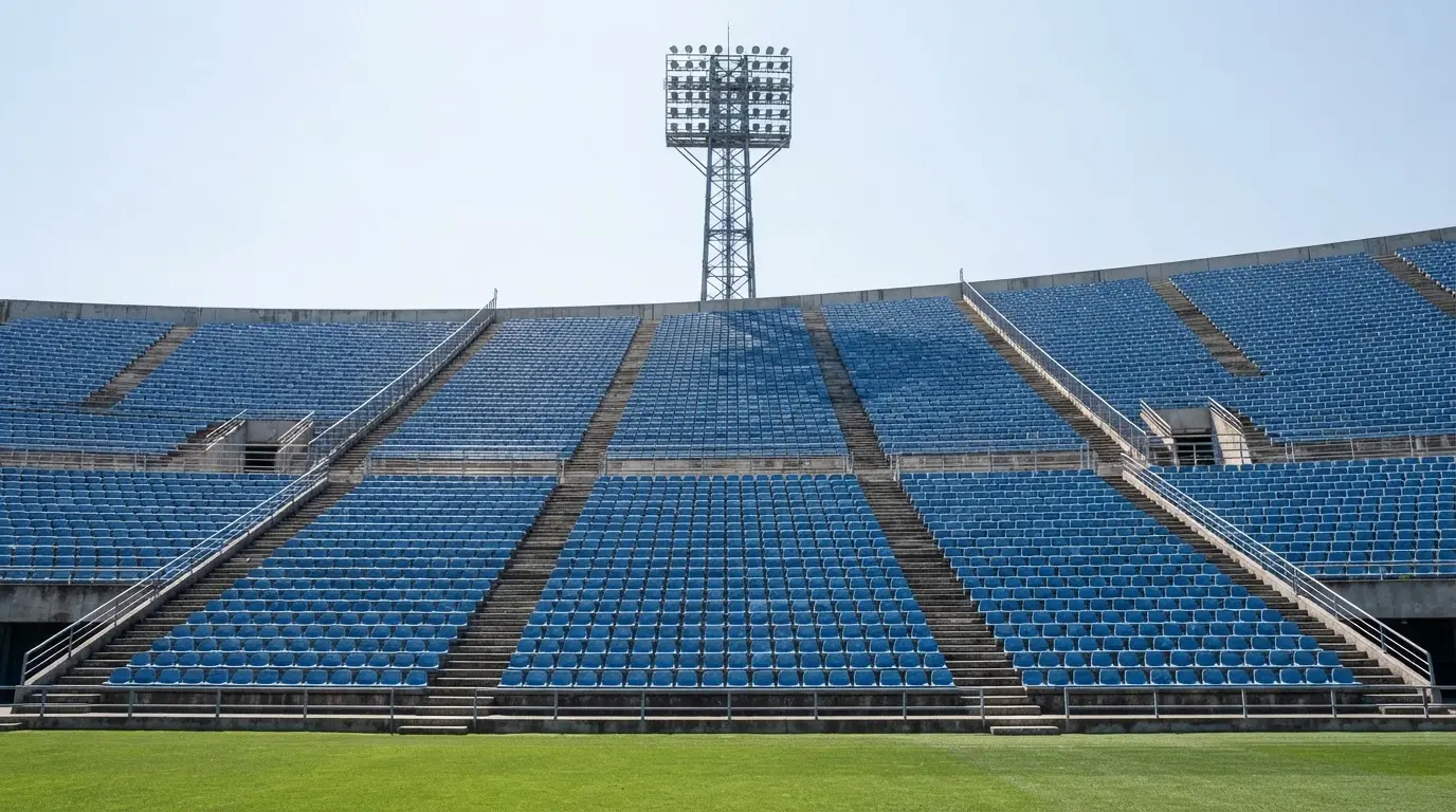 Eine leere Tribüne eines Fußballstadions bei Tageslicht, Sitzschalen in Reihen und dahinter ein Flutlichtmast.