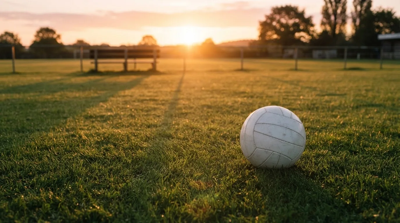 Ein Fußball auf einem Rasenplatz im Gegenlicht der tiefstehenden Sonne, am Rand eine leere Ersatzbank.
