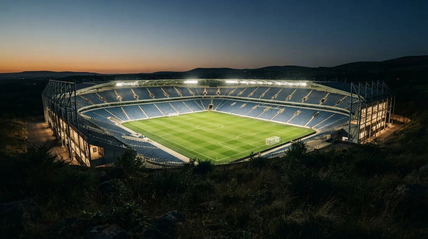 Ein leeres Stadion mit hell erleuchteten Rängen in der Abenddämmerung, von einem Hügel aus fotografiert.