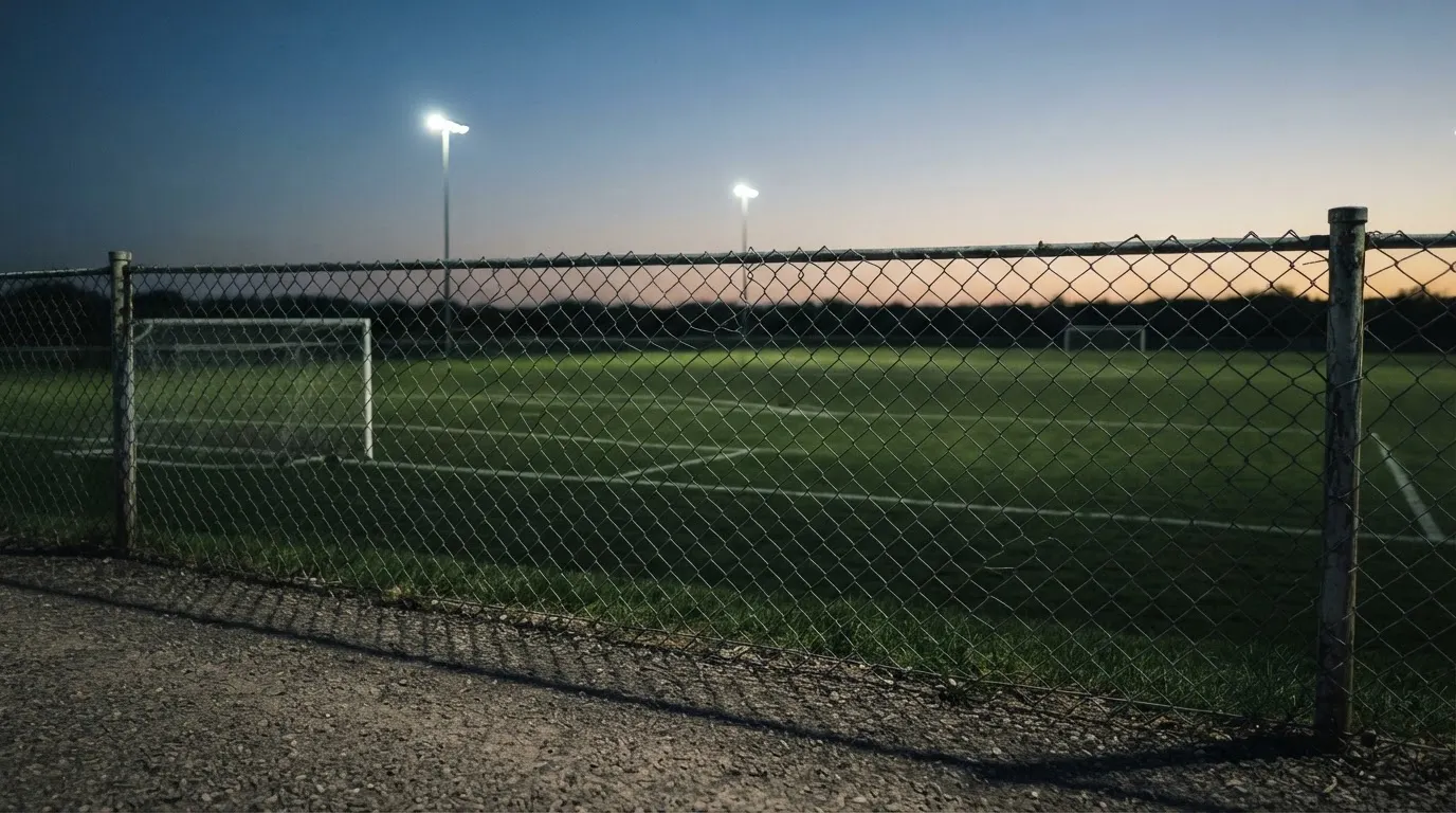 Eine Flutlichtanlage über einem leeren Sportplatz am Abend, im Vordergrund ein Maschendrahtzaun.