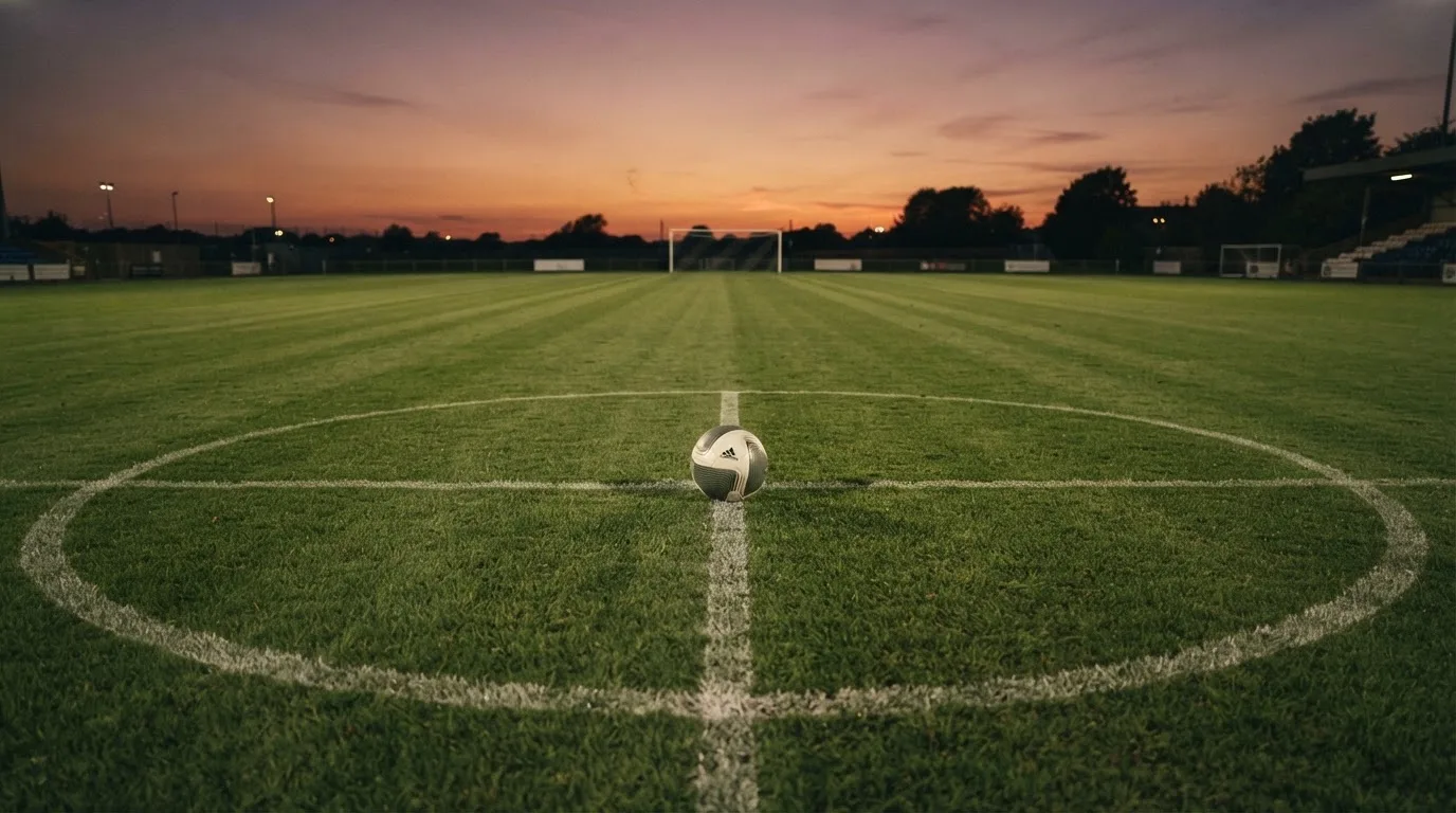 Ein Fußball liegt in der Mitte eines leeren Rasenplatzes, am Horizont färbt sich der Himmel orange.