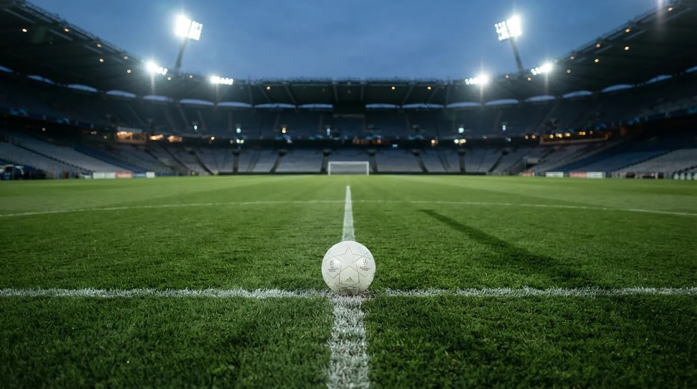 Ein Fußball liegt auf dem Anstoßpunkt eines leeren Rasenplatzes, im Hintergrund ein Stadion unter Flutlicht in der Abenddämmerung.