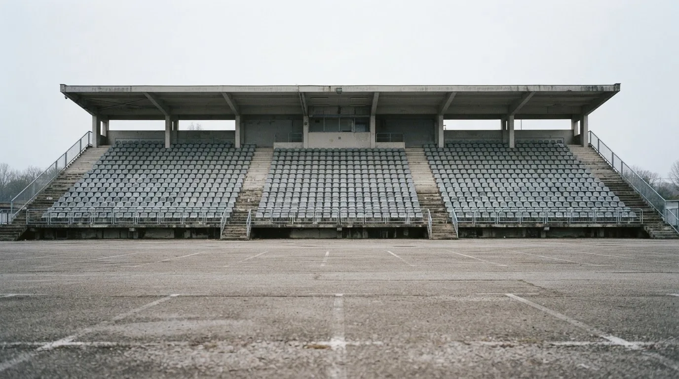 Eine leere Tribüne eines kleinen Stadions am frühen Morgen, aus der Ferne über einen menschenleeren Parkplatz fotografiert.