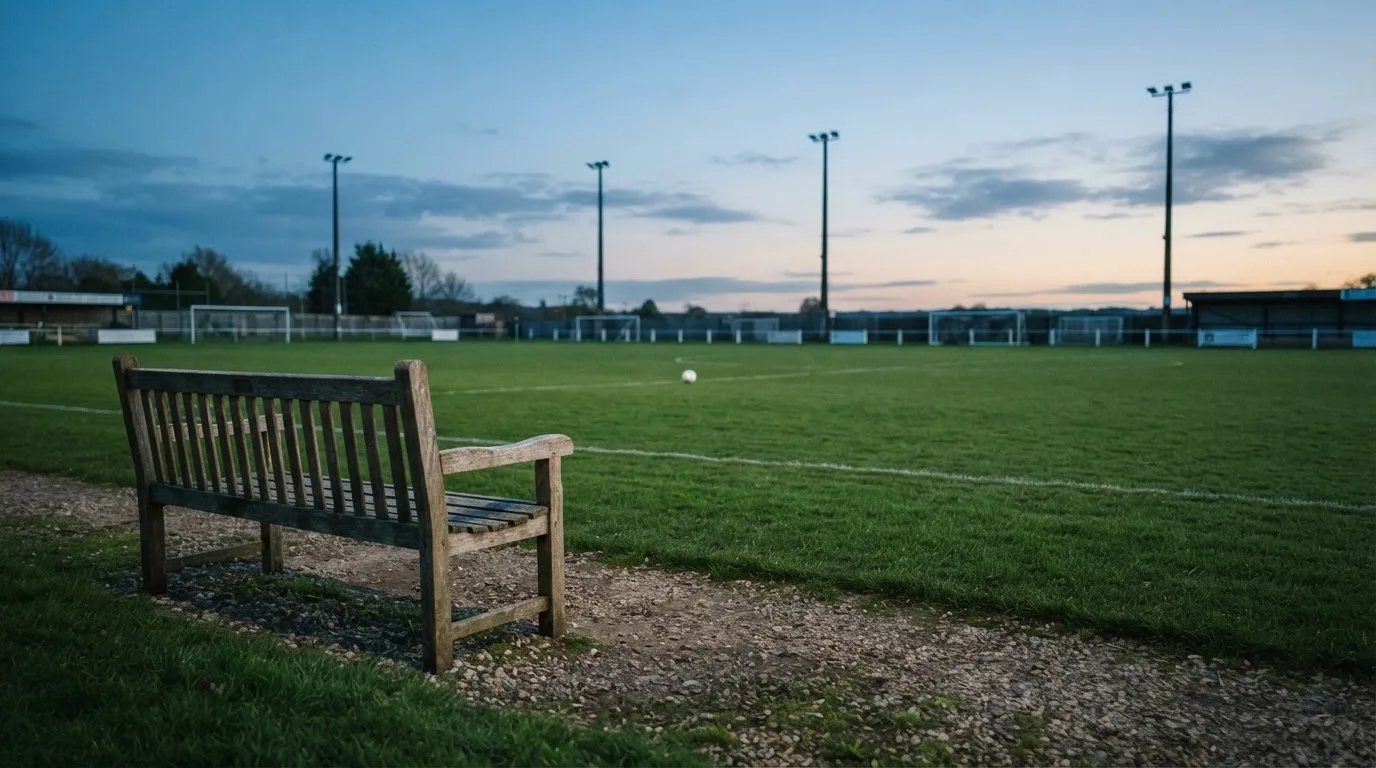 Eine leere Parkbank am Rand eines Fußballfelds in der Abenddämmerung, im Hintergrund ragen dunkle Flutlichtmasten in den Himmel.