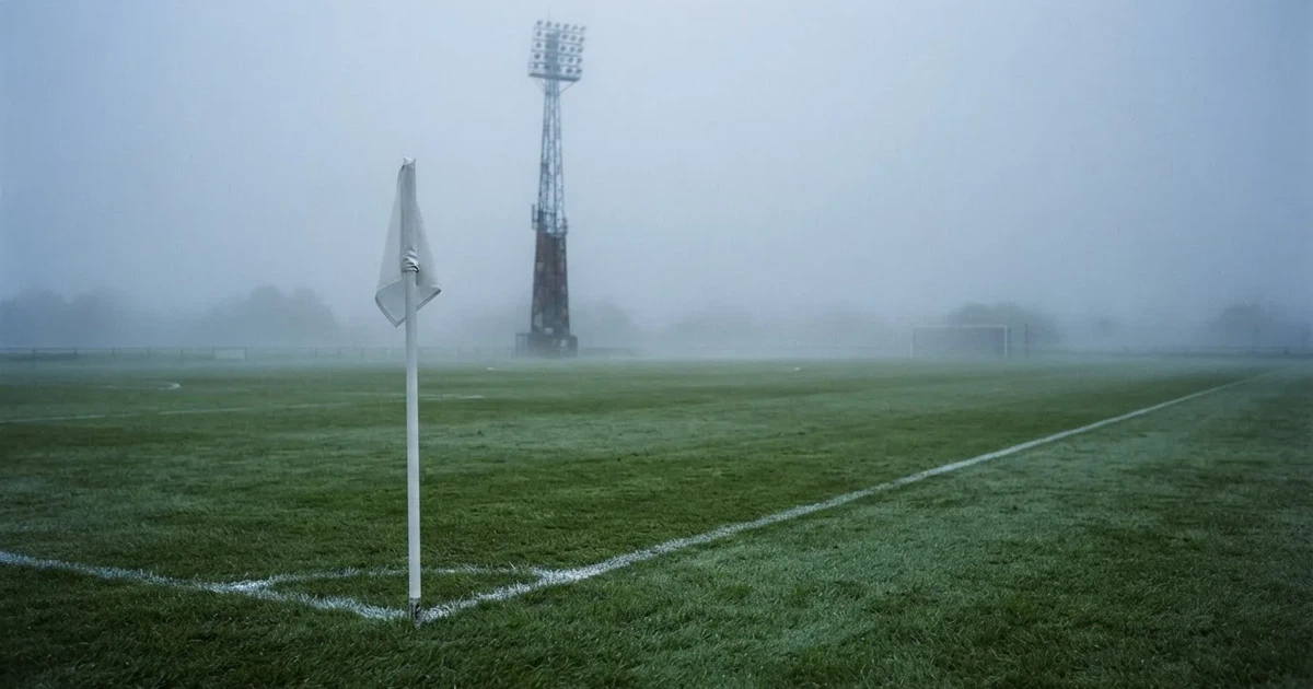 Eine Eckfahne steht im Nebel am Rand eines leeren Rasenplatzes in der Morgendämmerung, dahinter ein Flutlichtmast.