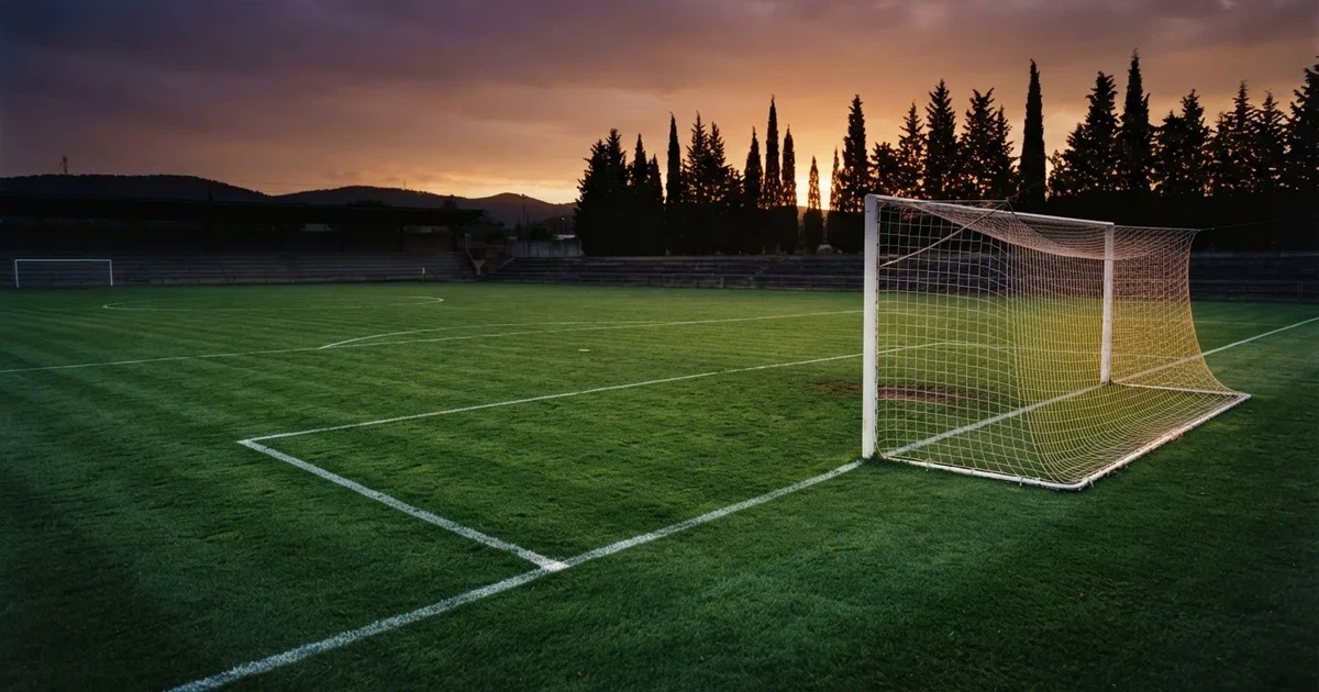 Ein Fußballtor mit Netz auf einem leeren Platz im Abendlicht, der Rasen frisch gemäht, im Hintergrund Zypressen.
