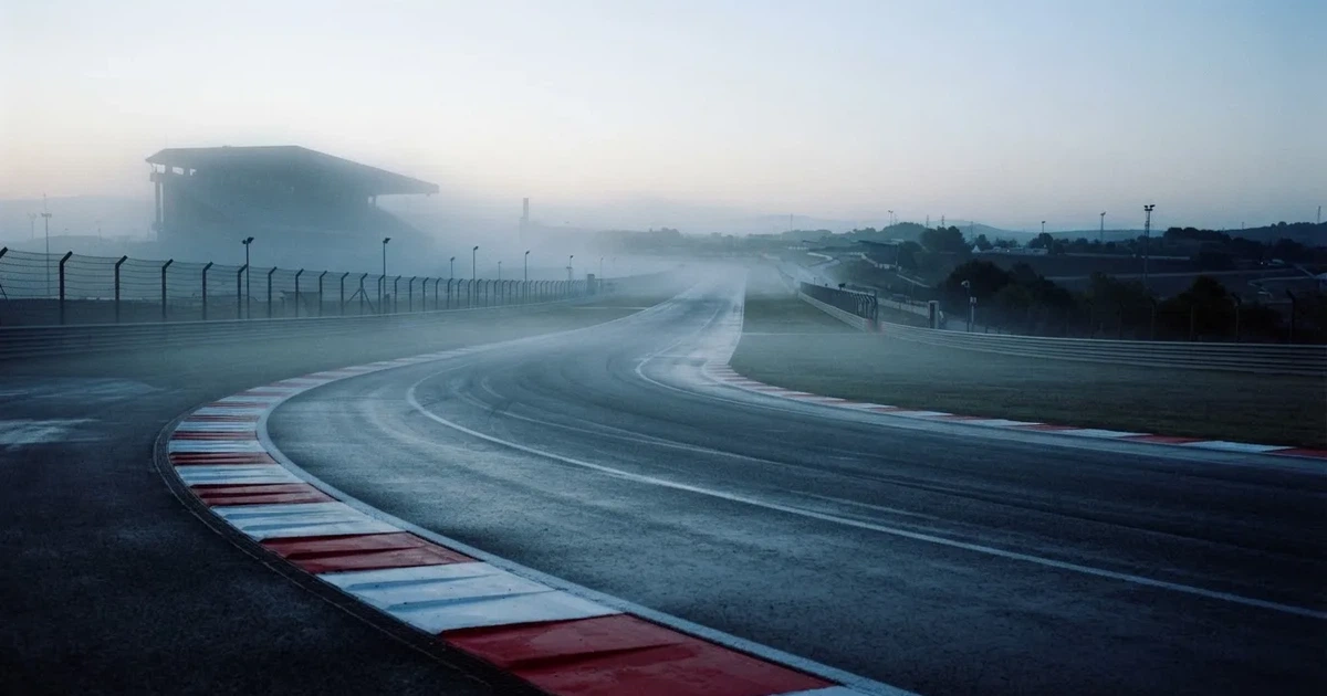 Eine leere Rennstrecke mit rot-weißen Randsteinen im frühen Morgenlicht, auf dem Asphalt liegt dünner Nebel, keine Fahrzeuge.