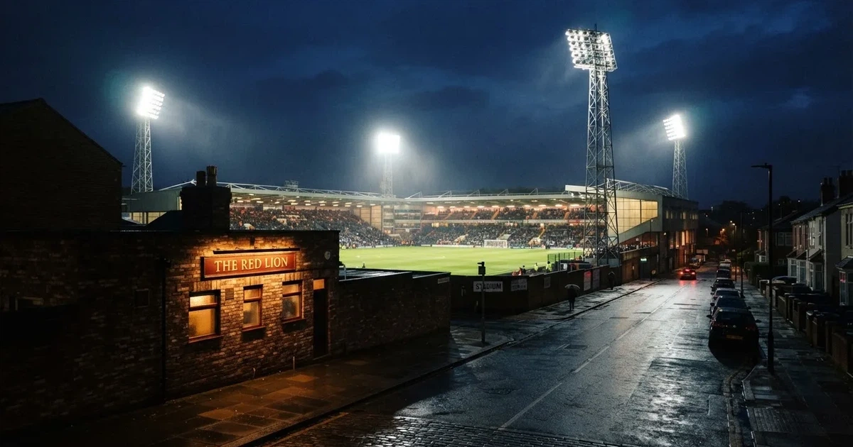 Ein Fußballstadion bei Flutlicht aus der Ferne gesehen, davor eine Straße und leuchtende Fenster eines Nebengebäudes.