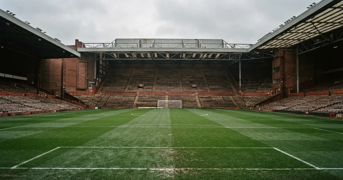 Ein englisches Fußballstadion mit rotbraunen Backsteintribünen unter bedecktem Himmel, davor ein nasser Rasen mit Spielfeldlinien.