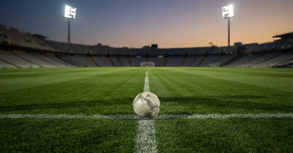 Ein Fußball liegt auf dem Anstoßpunkt eines leeren Rasenplatzes, im Hintergrund eine Tribüne und Flutlichtmasten in der Abenddämmerung.