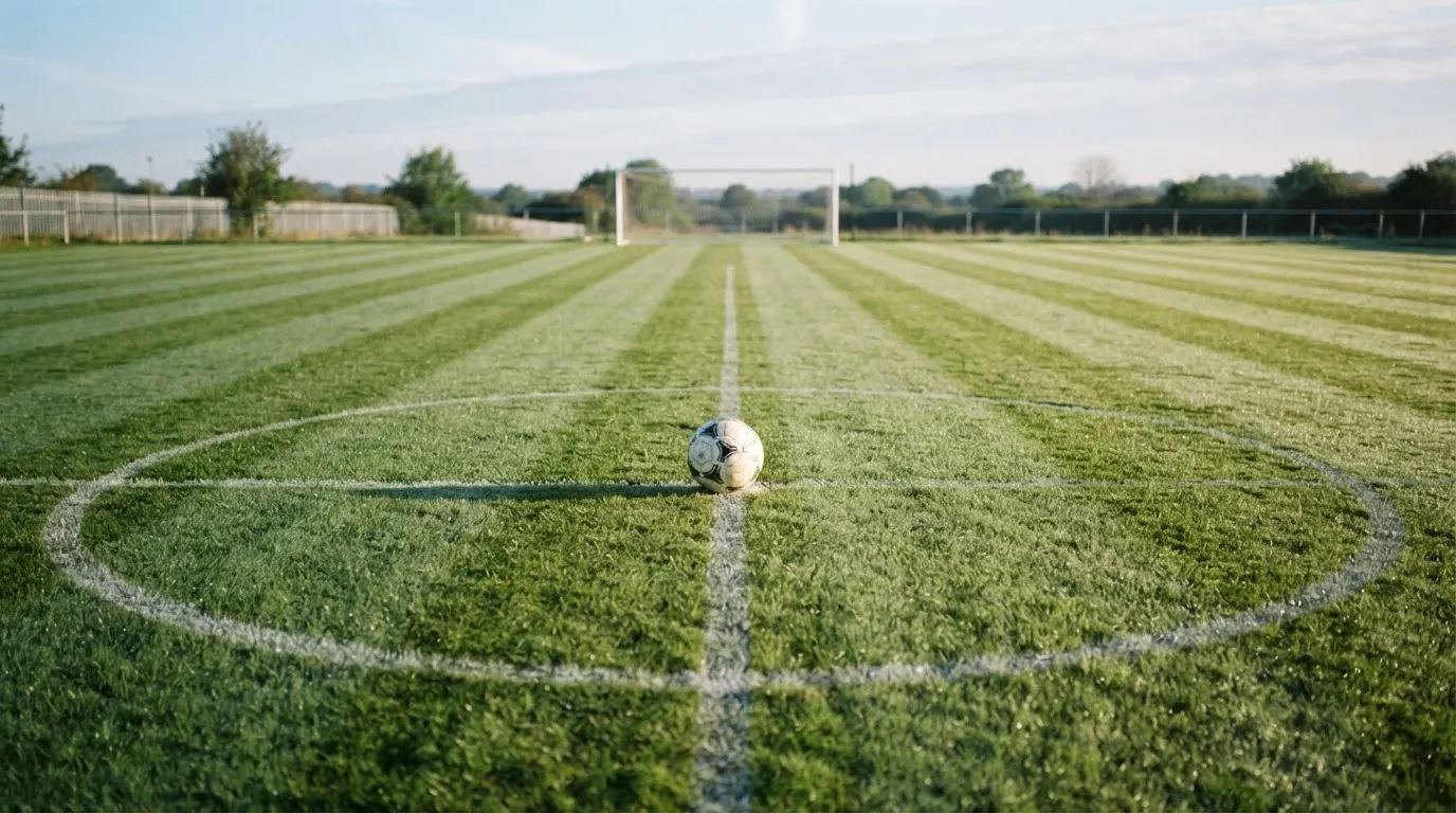 Ein Fußball liegt allein auf dem Anstoßpunkt eines leeren Rasenplatzes im Morgenlicht, am Rand ein niedriger Zaun.