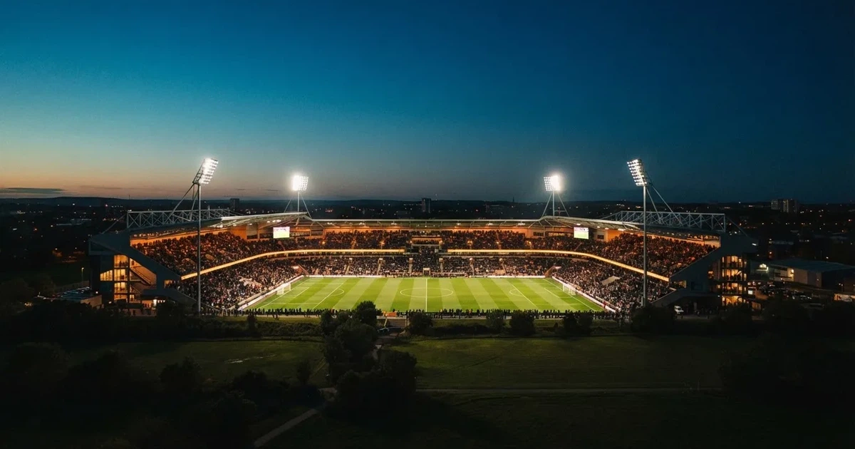 Ein Fußballstadion am Abend unter Flutlicht aus der Ferne gesehen, die Tribünen sind leicht beleuchtet und der Himmel dunkelblau.