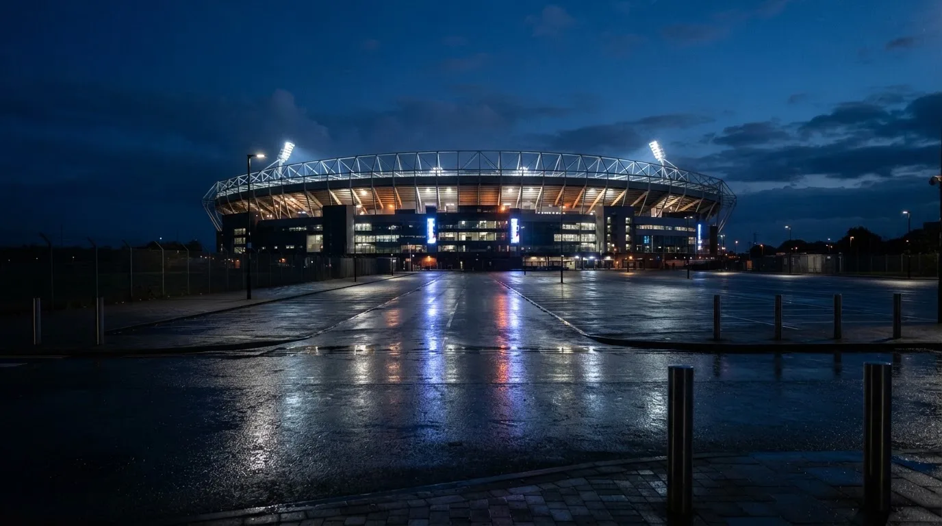 Ein Stadion unter Flutlicht von weitem betrachtet, davor ein stiller Platz bei Dunkelheit und feuchtem Asphalt.