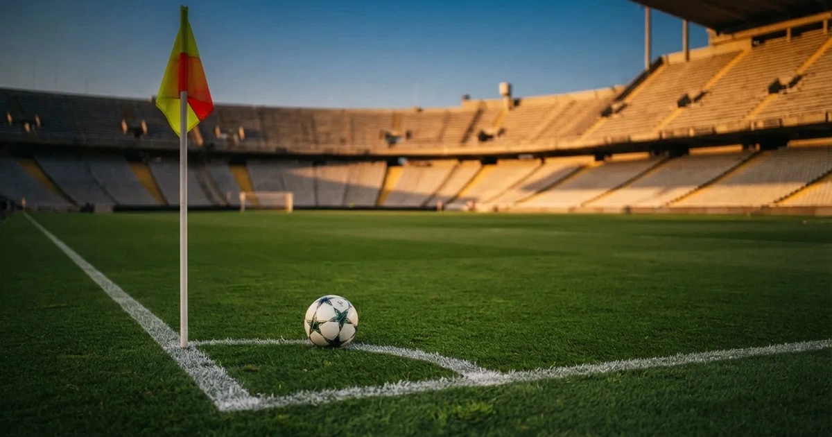 Ein Fußball liegt am Viertelkreis an der Eckfahne eines Rasenplatzes, im Hintergrund leere Tribünen in warmem Abendlicht.