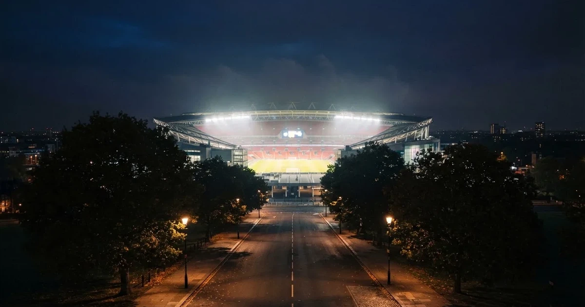 Ein Fußballstadion bei Nacht aus großer Entfernung, hell erleuchtet unter Flutlicht, davor eine dunkle Straße und Bäume.