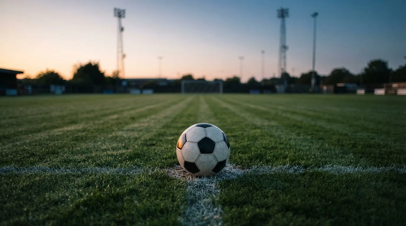 Ein Fußball liegt auf dem Anstoßpunkt eines Rasenplatzes in der Abenddämmerung, Flutlichtmasten stehen unscharf im Hintergrund.