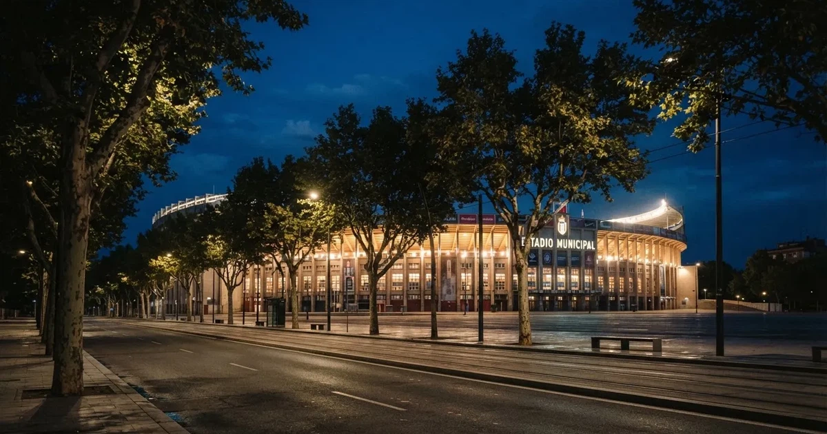 Ein hell erleuchtetes Stadion aus der Ferne bei Nacht, davor eine stille Straße und ein paar Bäume.