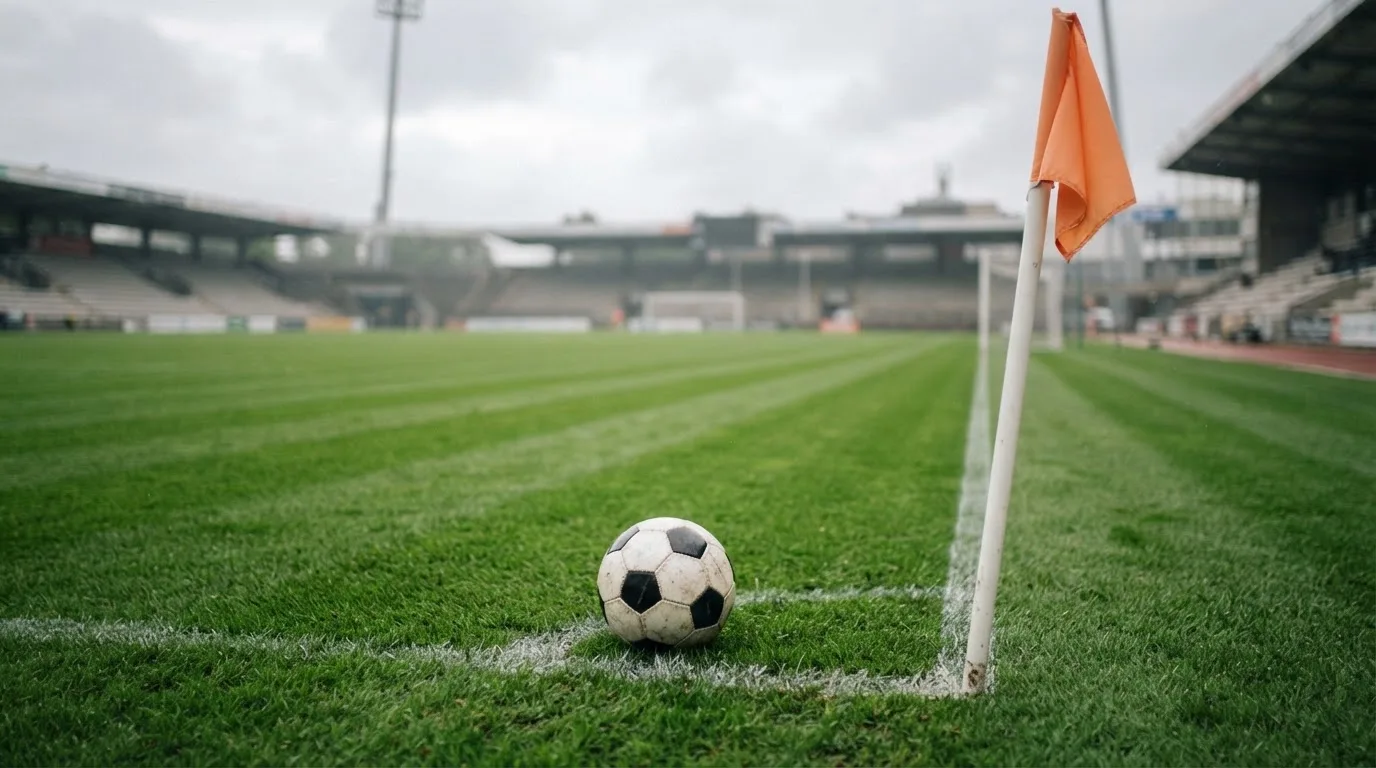 Ein Fußball liegt neben einer Eckfahne auf kurz gemähtem Rasen unter bewölktem Himmel.